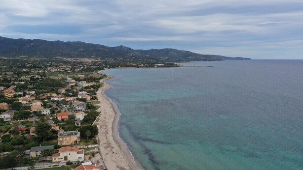 Vista aérea de un pueblo costero con una larga playa de arena, una zona residencial densa y montañas al fondo.
