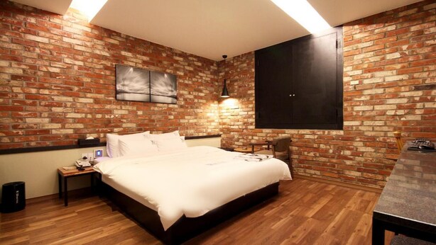 Un dormitorio con pared de ladrillo, cama, mesita de noche, lámpara y televisor montado en la pared.