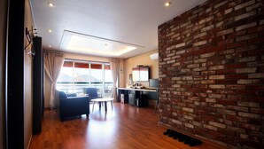 Una habitación de hotel moderna con piso de madera, una pared de acento de ladrillo, un ventanal con cortinas, un área de estar con sofá y sillones, una mesita, un escritorio con computadora y un televisor.