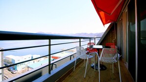 Un balcón con vistas al agua y a las montañas, con una mesa y sillas bajo un paraguas rojo.