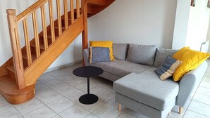 A modern living room with a wooden staircase, grey sofa, and a small round table. - Beachside House with Garden & WiFi, 400m from Shore in Pleumeur-Bodou (Pleumeur-Bodou)