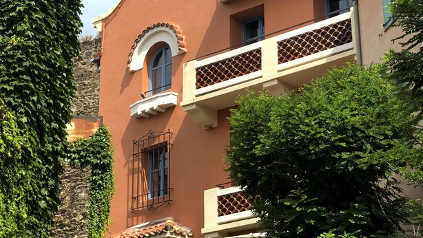 Ein Gebäude mit Balkon, einem Fenster mit dekorativem Bogen und einer Steinmauer, die teilweise von Efeu bedeckt ist.