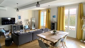 Ein modernes Wohnzimmer mit einer Sofagarnitur, einem Esstisch und Stühlen. An der Wand ist ein Fernseher angebracht, es gibt eine Lampe und ein Fenster mit Vorhängen.
