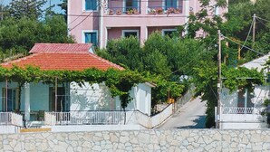 A pink multi-story building with balconies, a stone wall, and a red-tiled roof. - Natassa Apartments, 2 Bedrooms 30m from sea (Kefalonia)