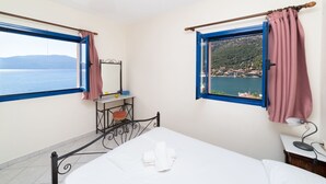 A bedroom with a bed, a bedside table, a lamp, a small table with a mirror, and a view of the sea and hills. - Natassa Apartments, 2 Bedrooms 30m from sea (Kefalonia)
