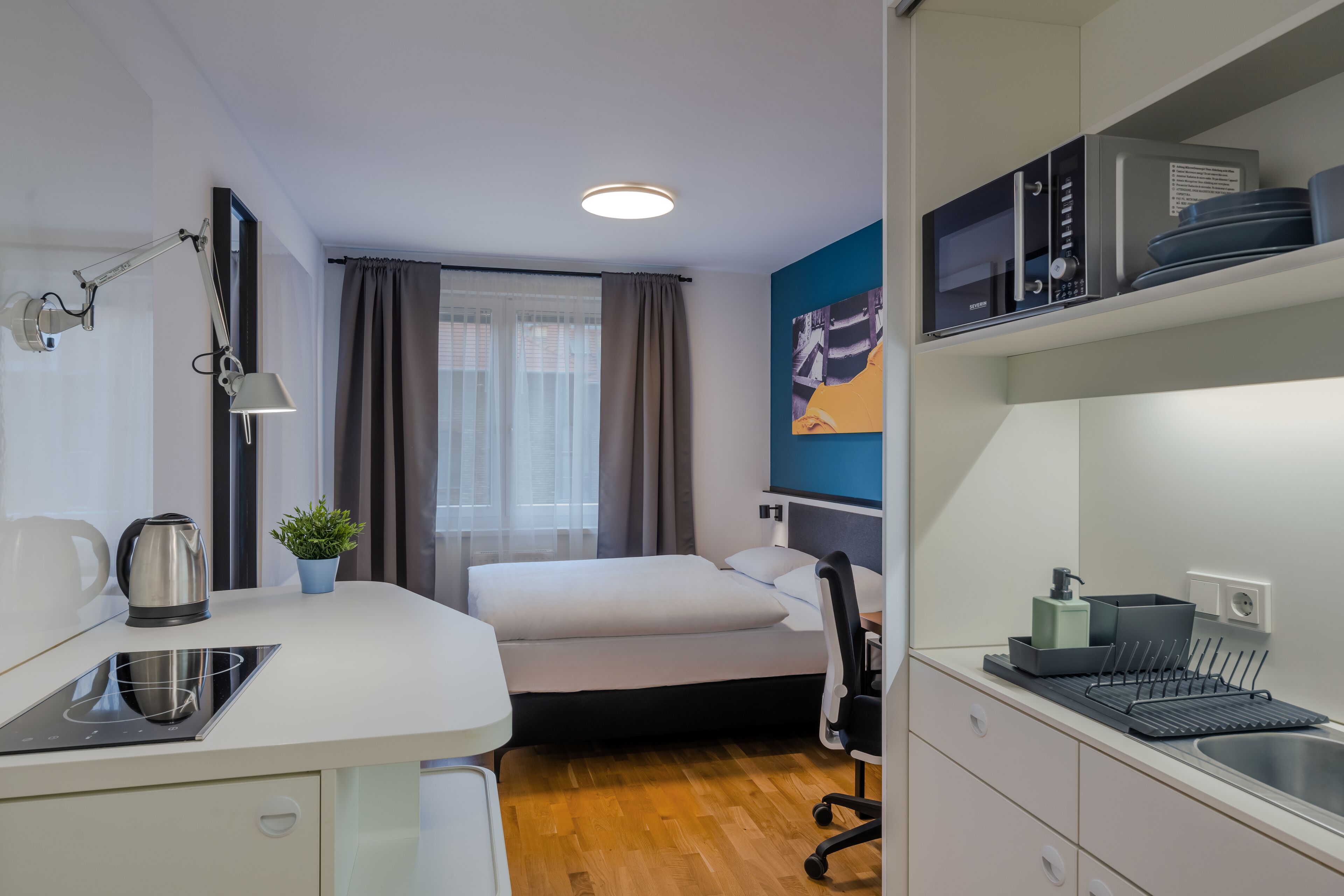 Ett kompakt hotellrum med ett litet pentry, en säng, ett skrivbord och ett fönster med gardiner.