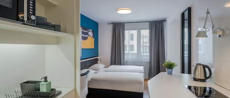 A modern hotel room with a bed, bedside table, and a small kitchenette.