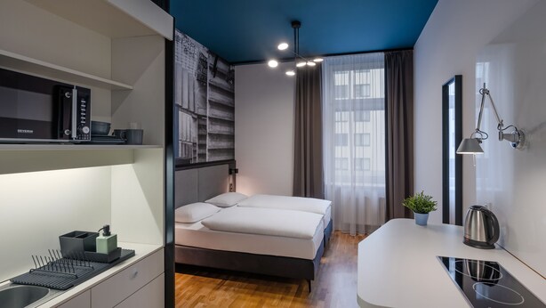 Un dormitorio moderno con cama, mesita de noche y una zona de cocina integrada que incluye microondas y hervidor.