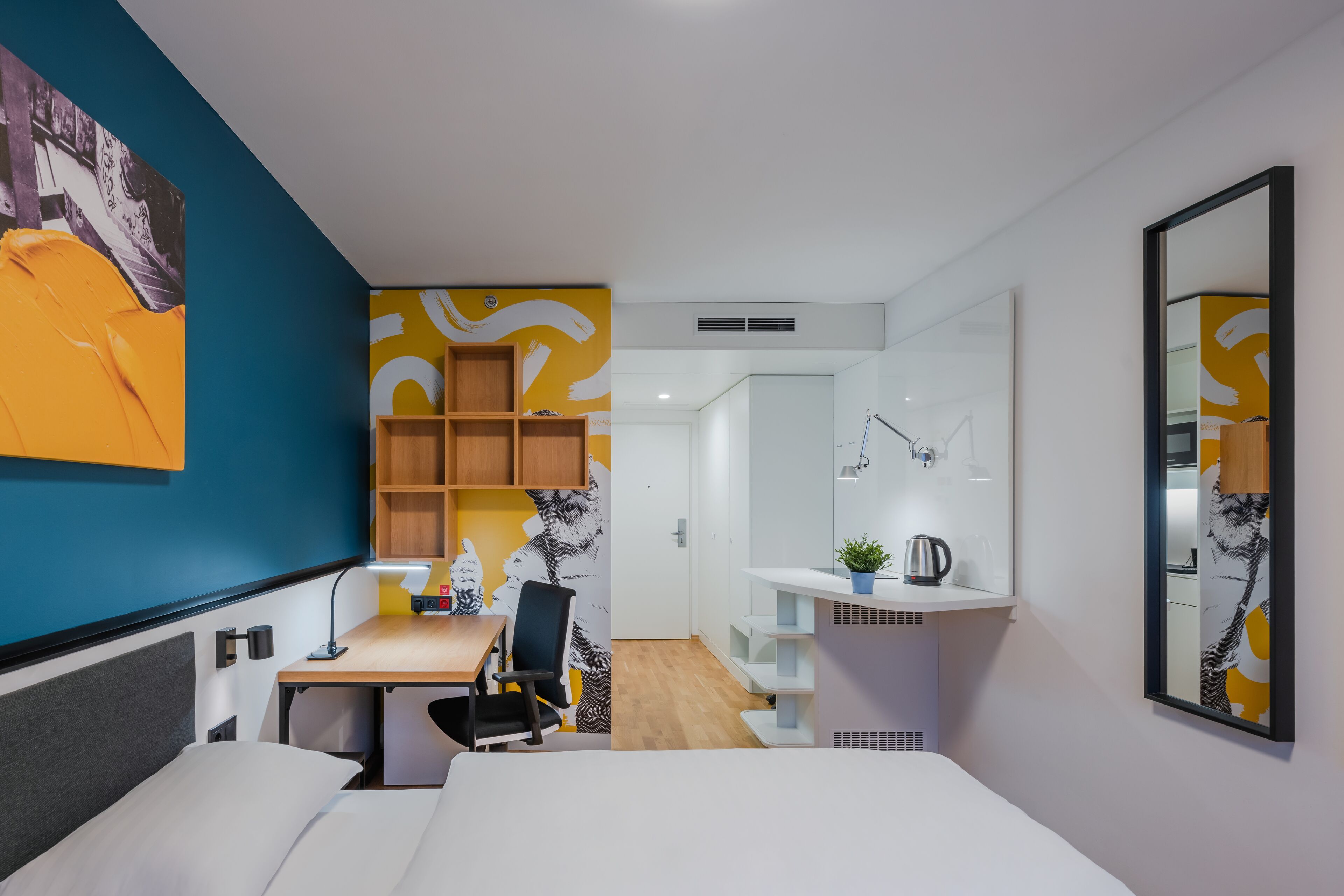 A modern hotel room with a bed, desk, chair, and a small kitchenette area.