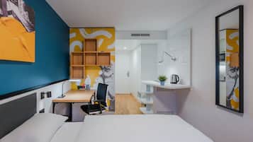 A modern hotel room with a bed, desk, chair, and a small kitchenette area.