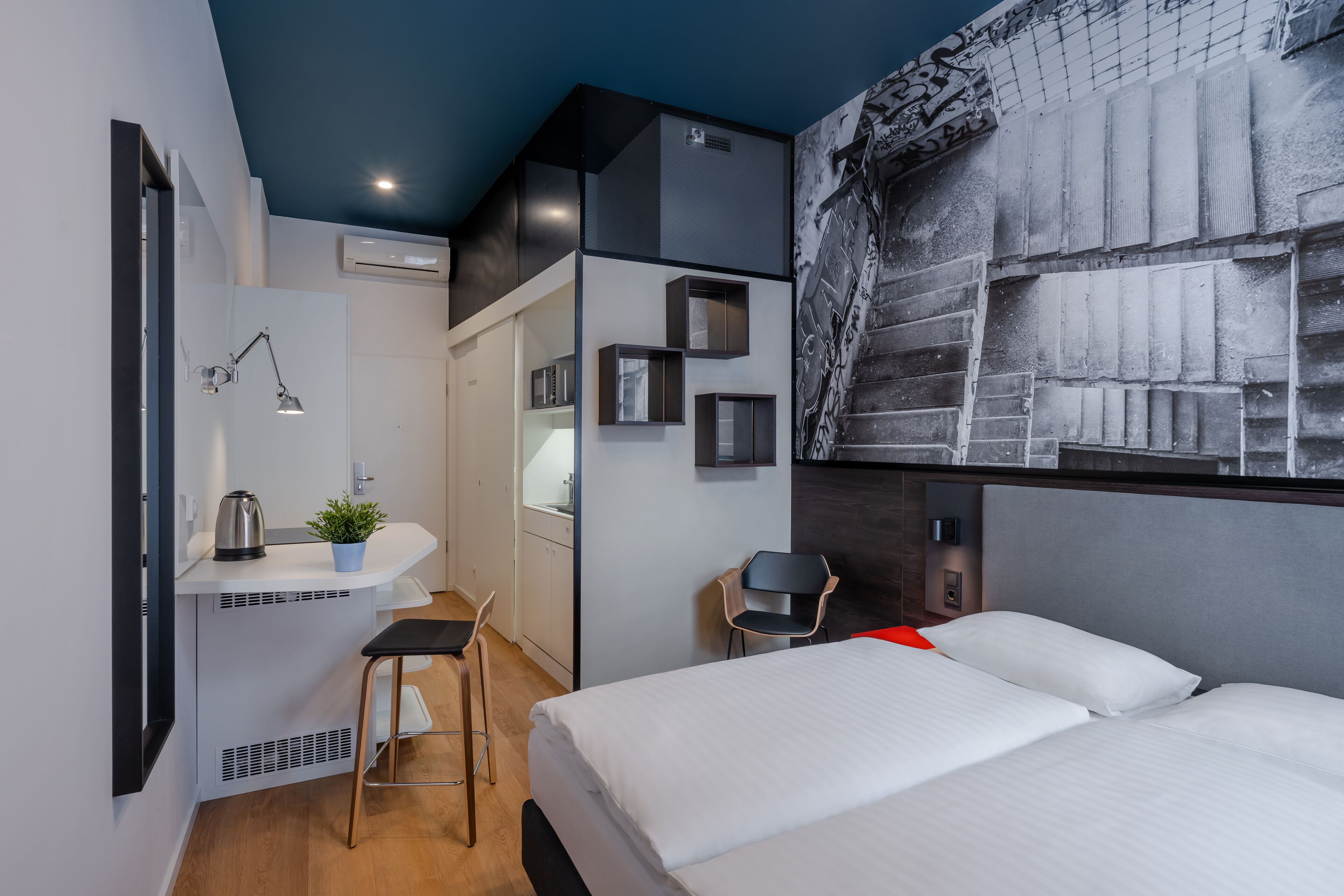 Een moderne hotelkamer met een bed, een bureau met stoel, een klein krukje, en een wandplank met een plant en een waterkoker.