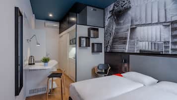 Een moderne hotelkamer met een bed, een bureau met stoel, een klein krukje, en een wandplank met een plant en een waterkoker.
