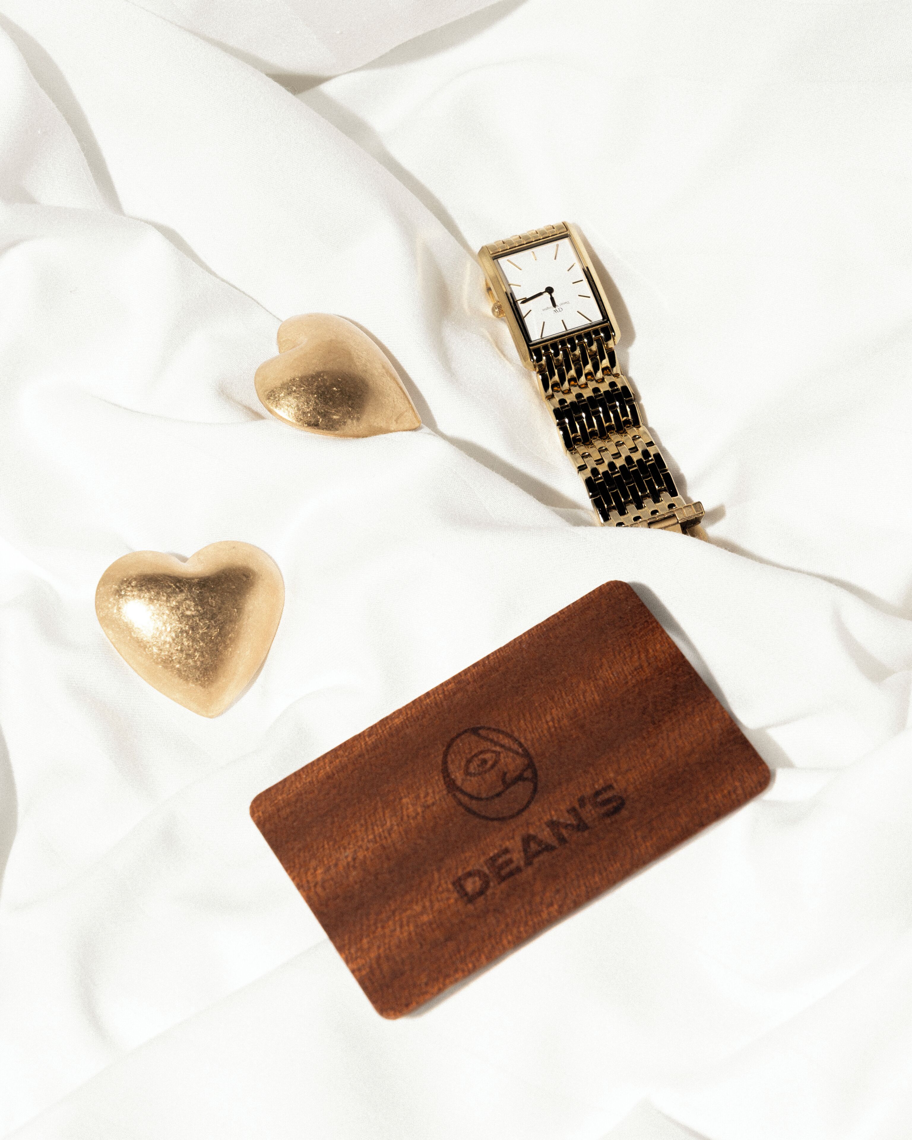 Una superficie blanca de tela con un reloj, un objeto con forma de corazón y una etiqueta de cuero con la palabra "DEANS" grabada.