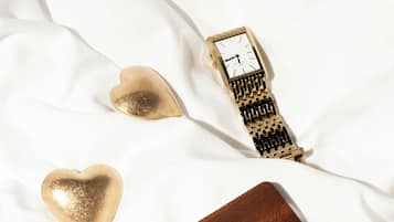 Una superficie blanca de tela con un reloj, un objeto con forma de corazón y una etiqueta de cuero con la palabra "DEANS" grabada.