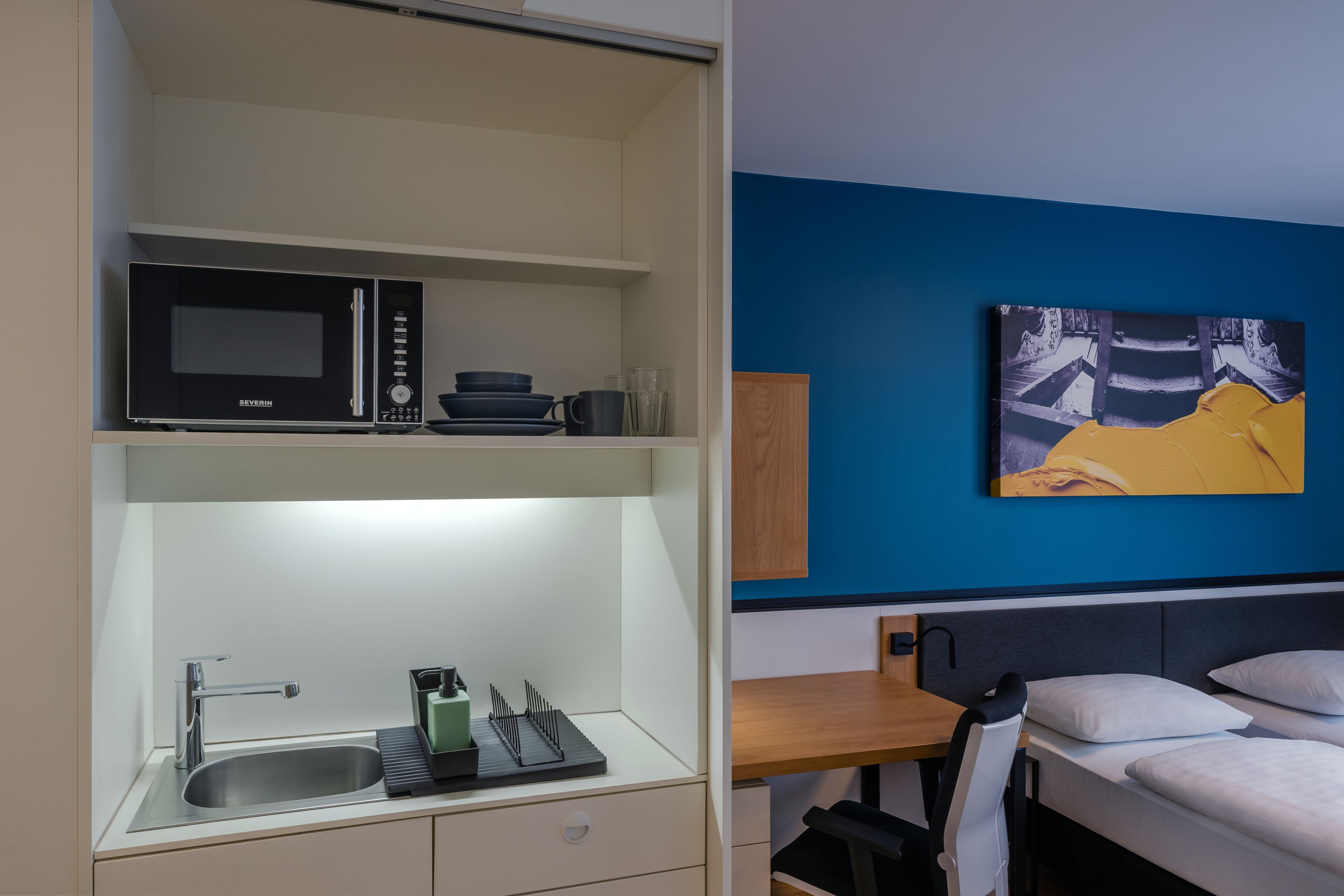 A compact hotel room with a kitchenette, a bed, a desk, and a wall-mounted painting.