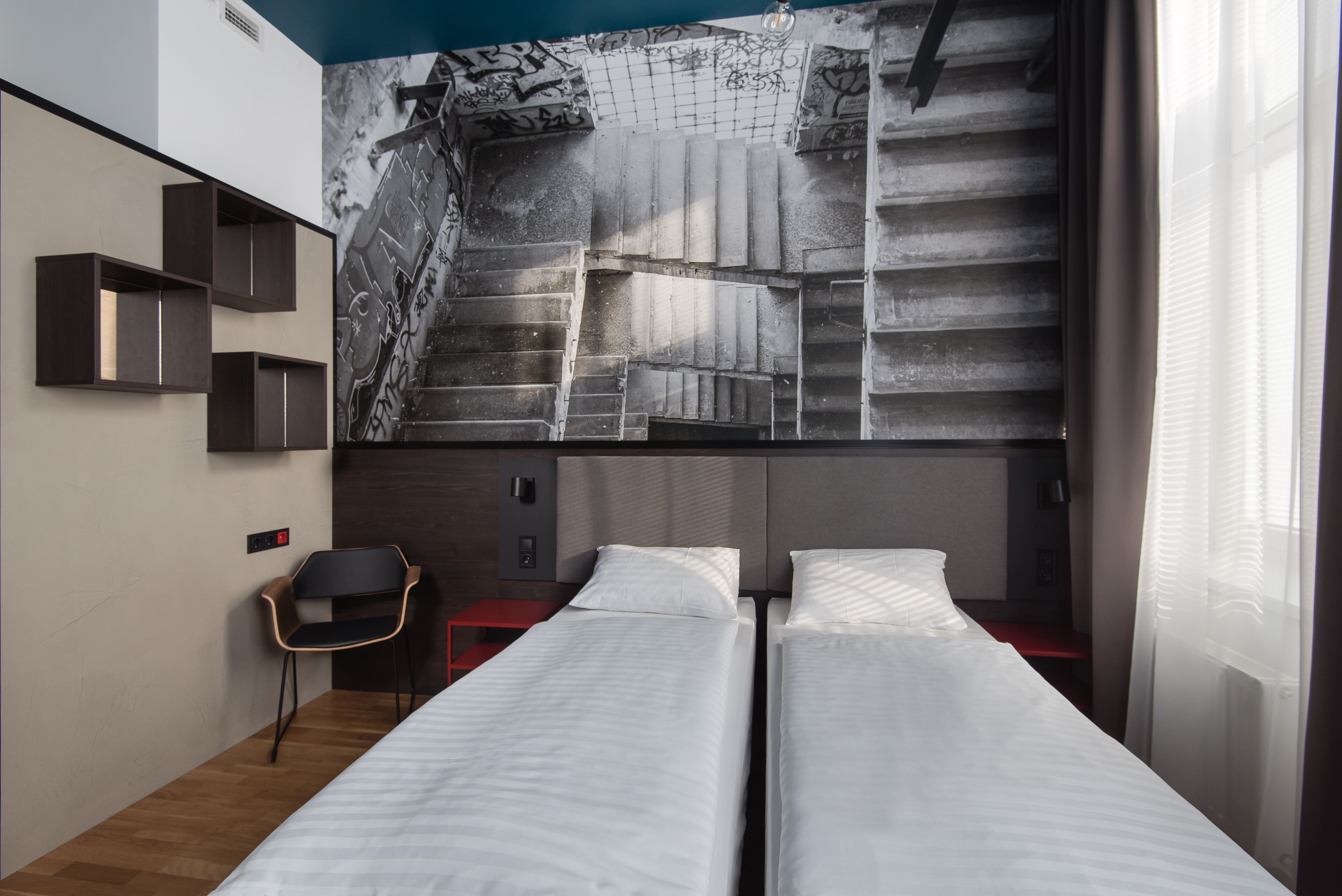 A hotel room with two beds, a mural on the wall, a chair, and a red nightstand.