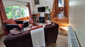 Ein gemütliches Wohnzimmer mit einer Couch, einem Fernseher, einem Bücherregal und einem Fenster mit Blick auf Grünflächen.