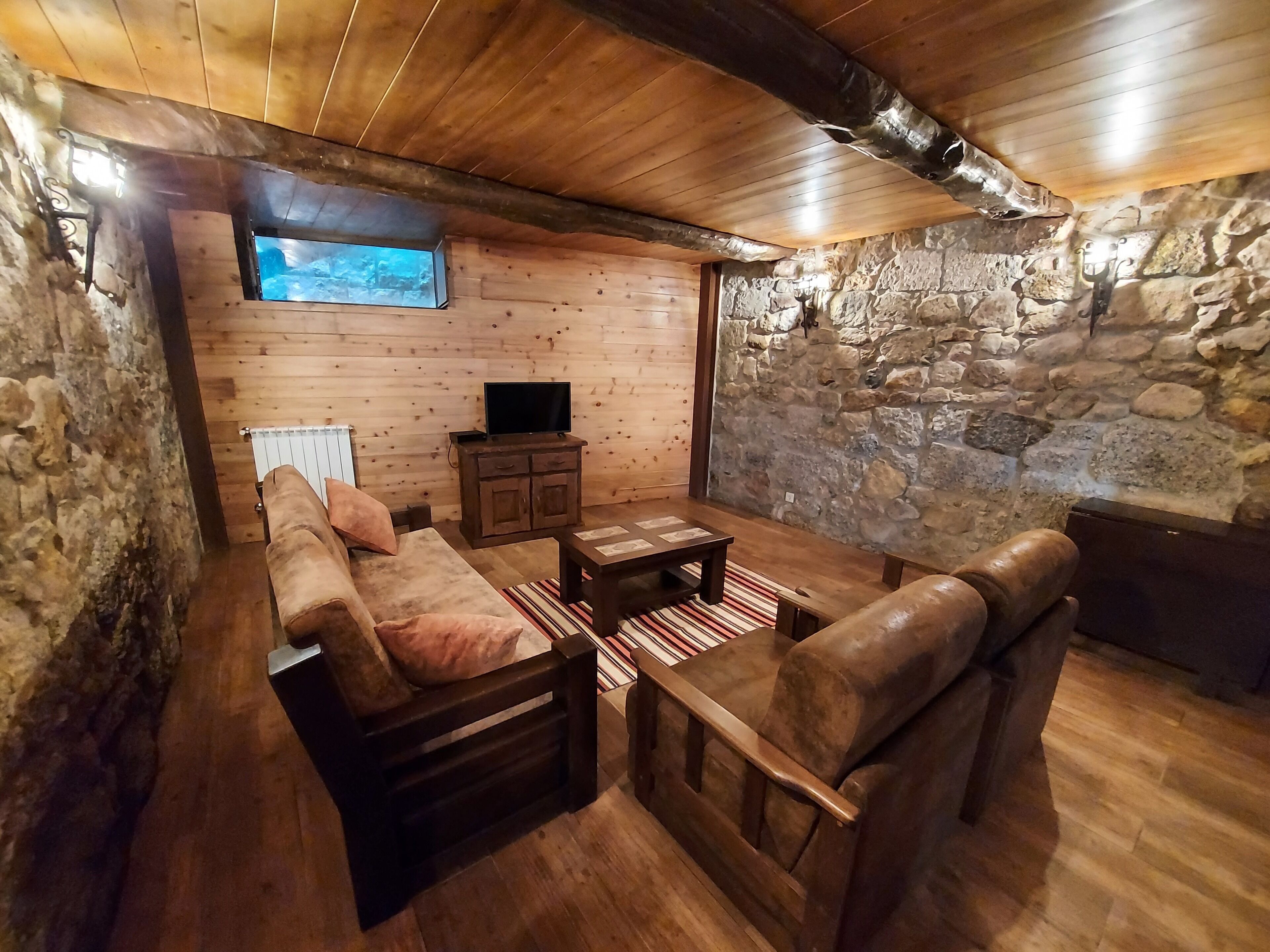 Una habitación acogedora con pared de piedra, techo de madera, un sofá, una silla, una mesita y un televisor sobre un mueble.