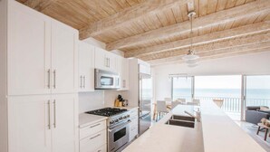Een moderne keuken met witte kasten, een ingebouwde oven en een magnetron. Er is een eethoek met een tafel en stoelen, en een uitzicht op de oceaan door grote ramen.