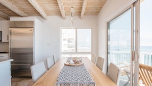 Een eethoek met een houten tafel, stoelen en een macramé tafelkleed. Grote ramen bieden uitzicht op de oceaan.