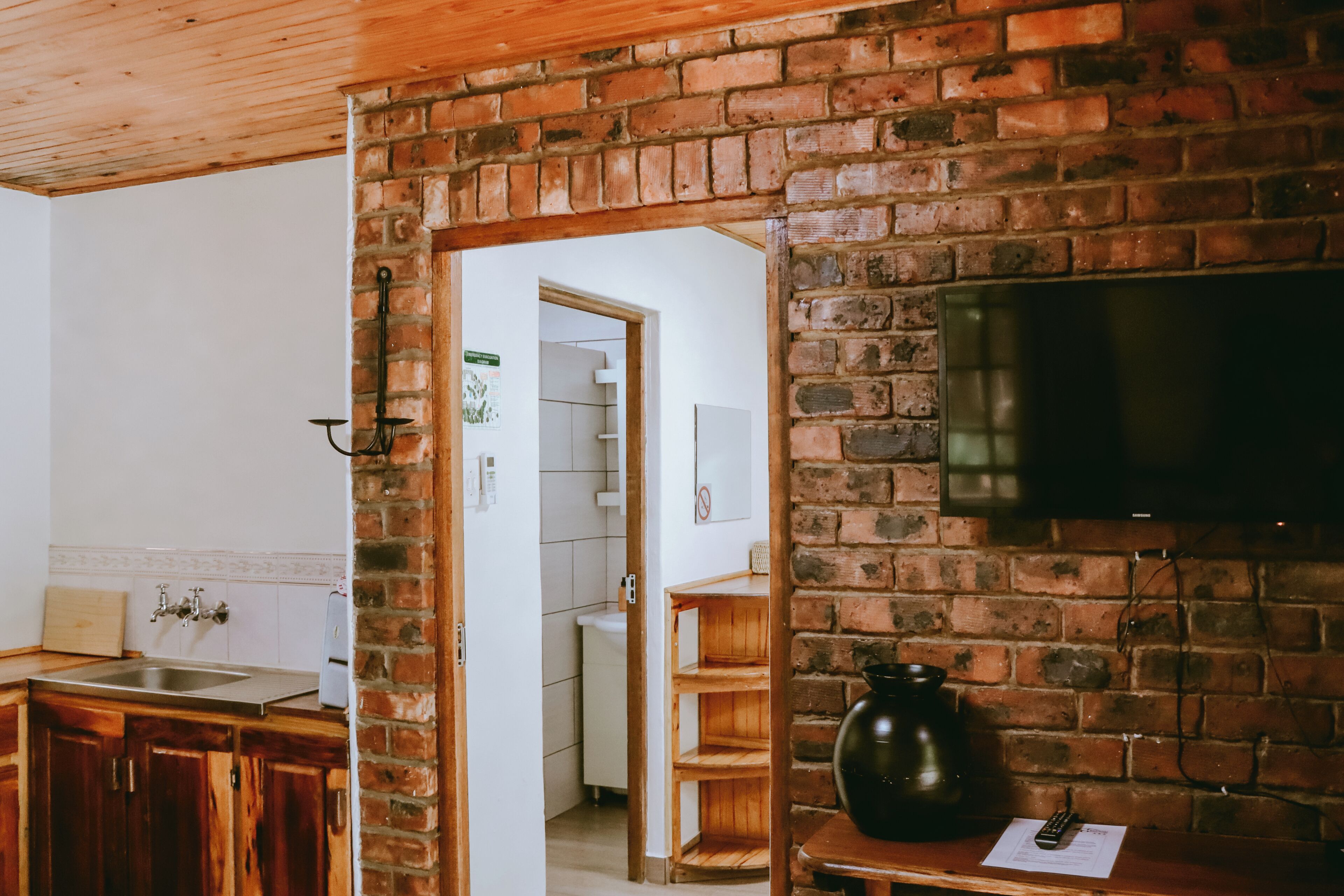 A kitchen with wooden cabinets, a brick wall, a TV mounted on the wall, and a wooden shelf with a vase.
