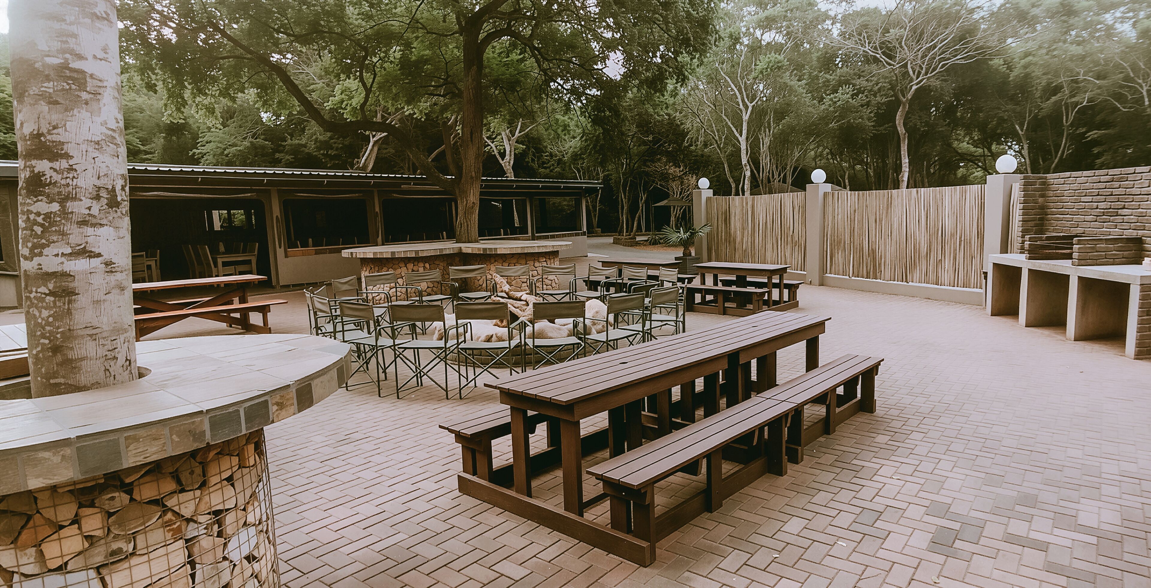 An outdoor patio area with wooden benches and tables, surrounded by trees and a stone wall.