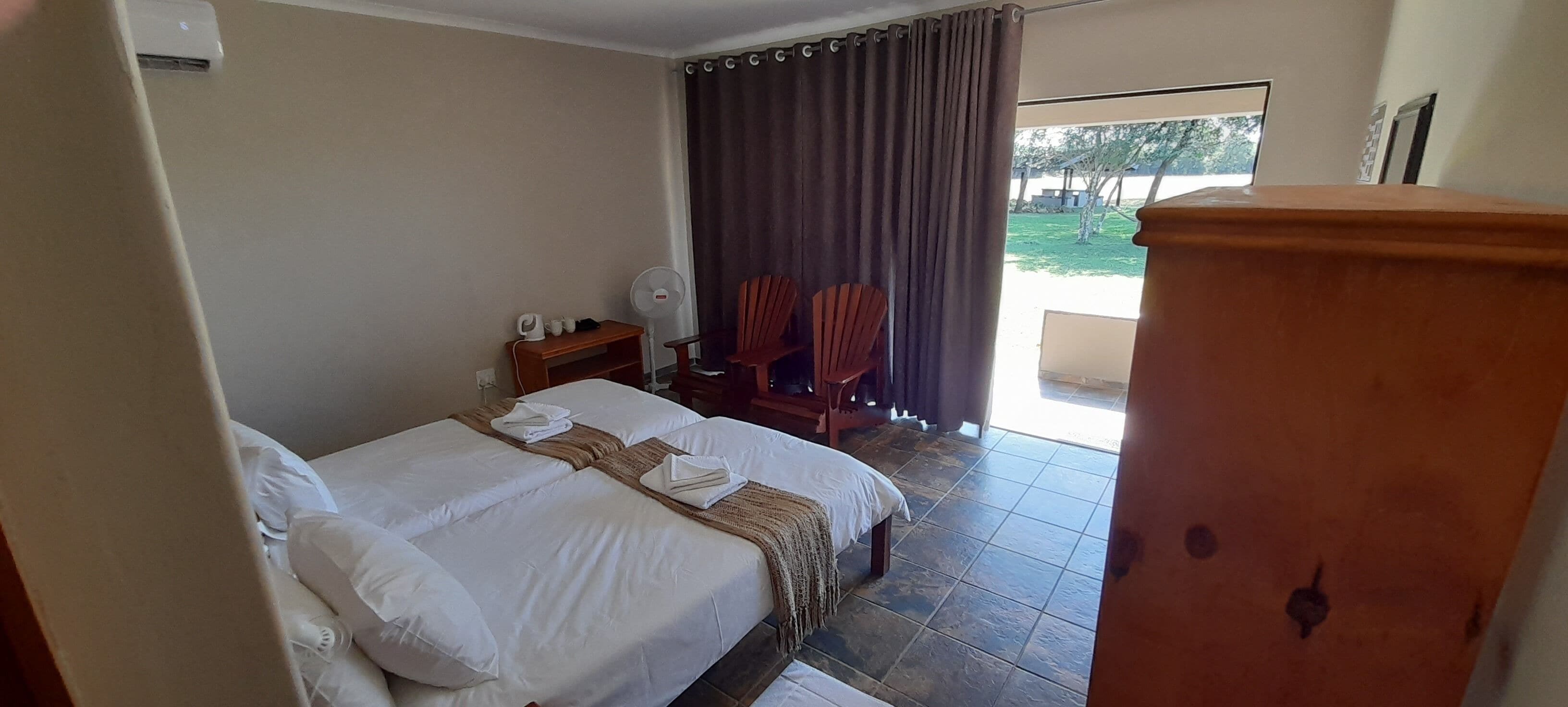 A hotel room with a bed, a wooden nightstand, a fan, and two chairs.