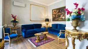 Una sala de estar con sofás azules, un tapete estampado, un cuadro con marco dorado y un jarrón con flores.