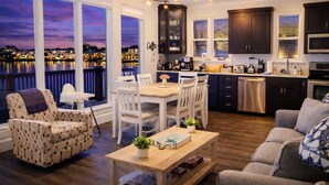 A modern kitchen with dark cabinets, a dining area with white chairs, and a view of a waterfront at dusk. - [Summer Deals on remaining dates] 3 min to Beach! Waterfront, Pool, Beach gear (Destin)