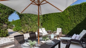 Un patio al aire libre con una mesa lista para comer, rodeado de sillones de descanso y una sombrilla grande.