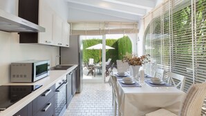 Una cocina moderna con microondas integrado, horno y armarios de acero inoxidable. Hay un comedor con una mesa preparada para dos, una silla blanca y una silla de mimbre. La habitación tiene un gran ventanal con persianas y vistas a un seto verde.