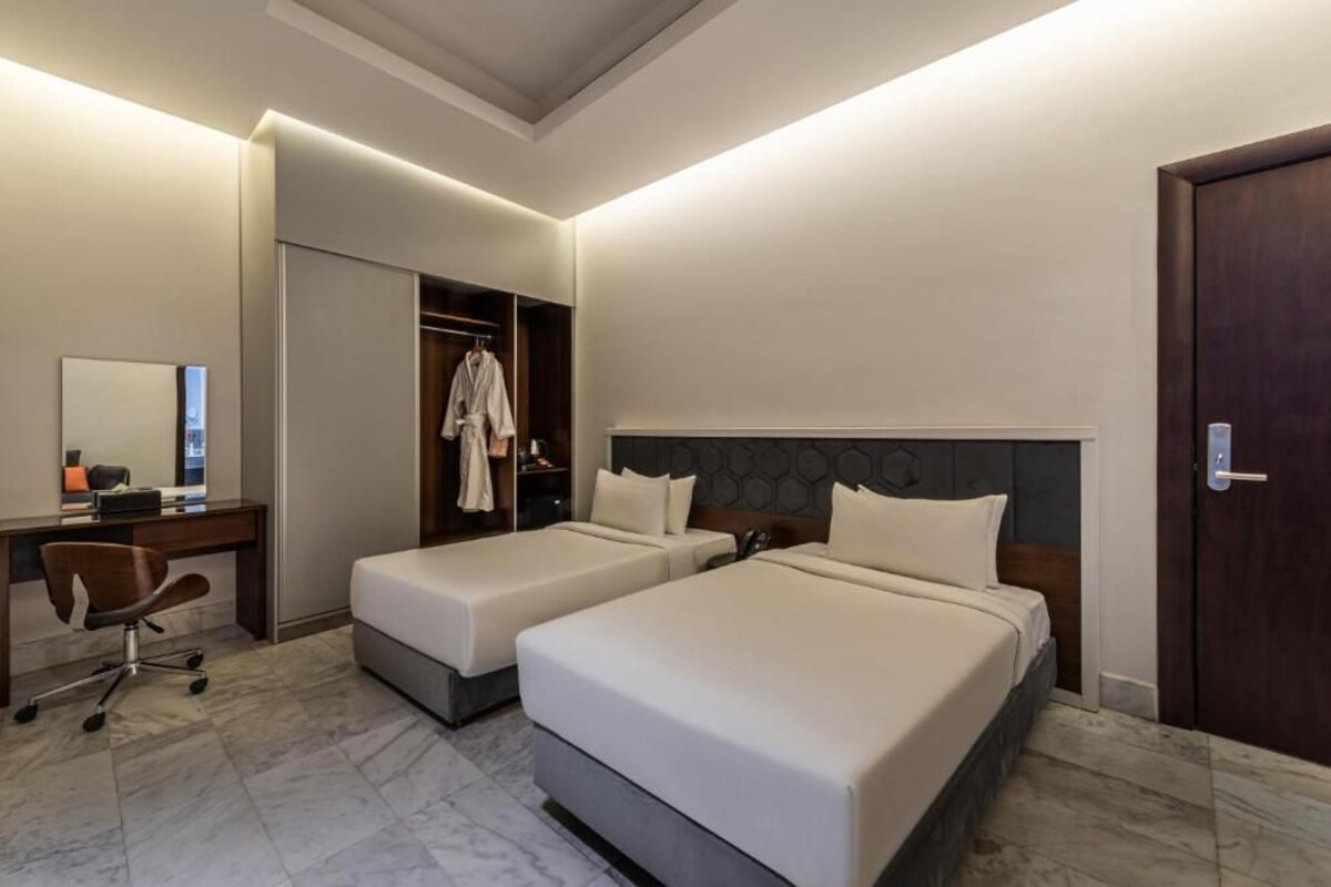 Quarto de hotel com duas camas, uma escrivaninha, uma cadeira, um guarda-roupa e um espelho.
