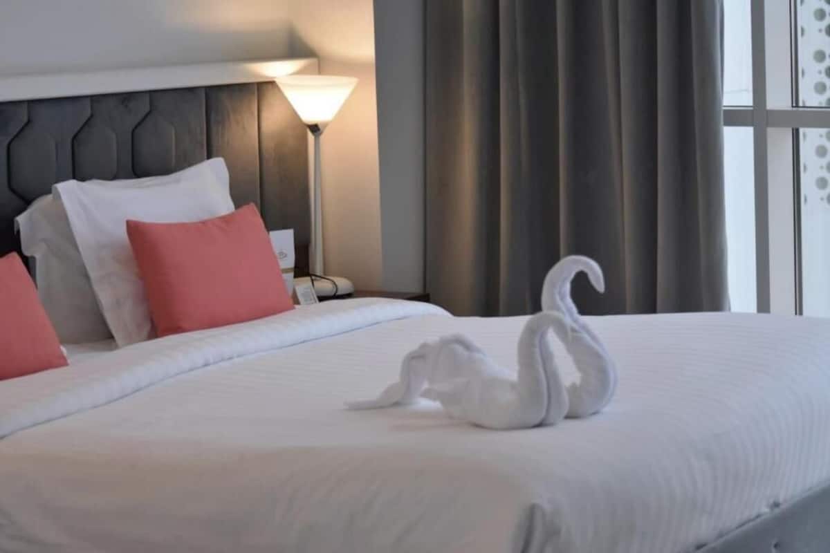 A neatly made bed with a swan-shaped towel, a lamp, and curtains in a hotel room.