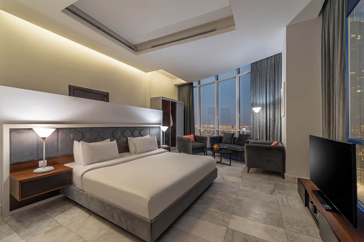 A modern hotel room with a large bed, a TV, a seating area with a view of the city, and a large window.