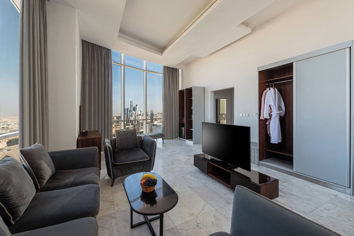 A modern hotel room with a city view, a flat-screen TV, a seating area with a coffee table, and a walk-in closet with robes.