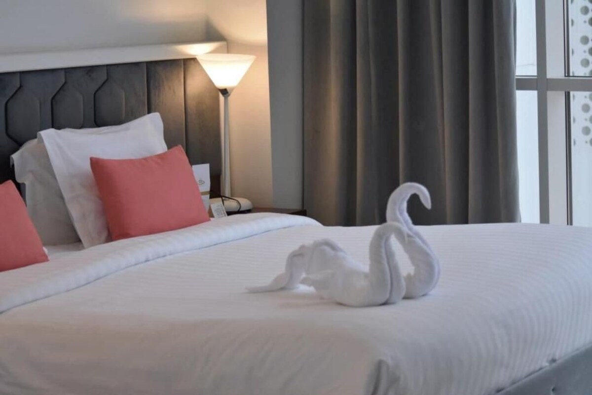 Cama bem arrumada com toalha em forma de cisne, luminária e cortinas em quarto de hotel.