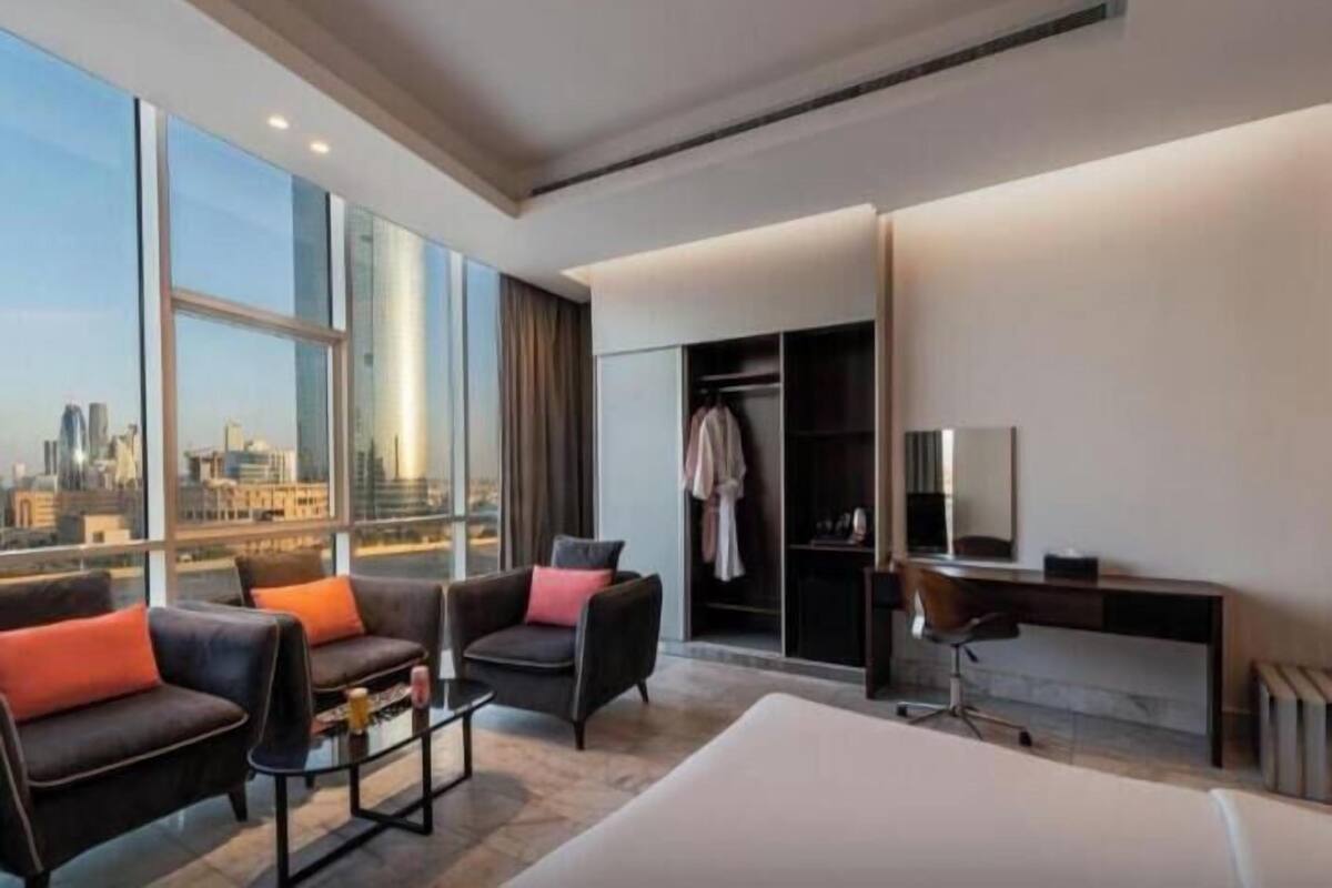 A modern hotel room with a large window, a bed, a desk, and a seating area.