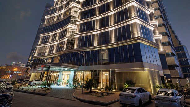 A modern high-rise building with a prominent central tower, multiple balconies, and a well-lit entrance area.