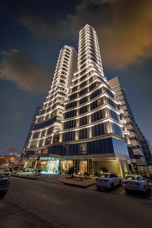 Un edificio moderno de gran altura con una torre central destacada, múltiples balcones y un área de entrada bien iluminada.