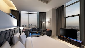 A modern hotel room with a large bed, a flat-screen TV, a seating area with a city view, and a balcony with a cityscape view.
