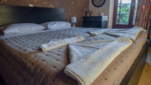 A neatly made bed with a tufted headboard, a bedside table with a lamp, and a view of greenery through a window. - Anthis Cottage (Malevizi)