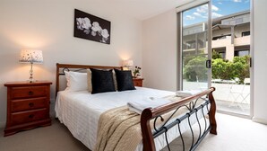 A bedroom with a wooden bed, white bedding, a dark wall art piece, a nightstand with a lamp, and a view of a balcony with outdoor furniture. - Blue Vista - a modern apartment (Gerringong)