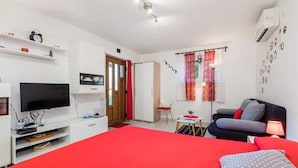 Ein Schlafzimmer mit roter Bettdecke, einem Fernseher, einem Schreibtisch mit Computer, einem Sessel und einem Sofa.
