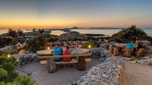 Ein schöner Essbereich im Freien mit Steintischen und -bänken, mit Blick auf eine Küstenlandschaft und einen Sonnenuntergang.