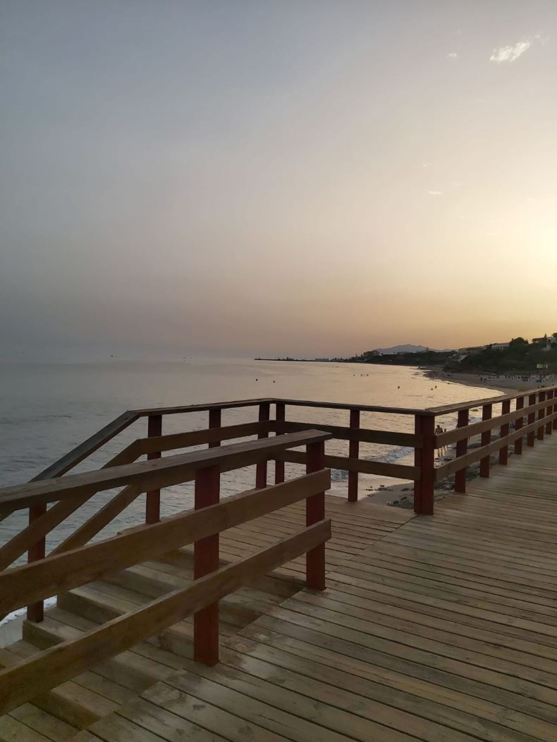 Une promenade en bois qui mène à une plage d’où l’on a une vue dégagée sur la mer et les collines lointaines.