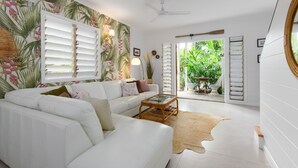 Un salon moderne avec un canapé blanc, une table basse et une vue sur un jardin luxuriant, visible à travers des portes ouvertes.