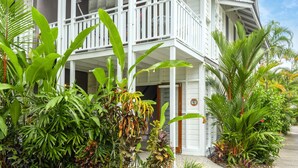 Un bâtiment de style colonial, peint en blanc, doté d’un balcon et d’une véranda, et entouré de plantes tropicales.