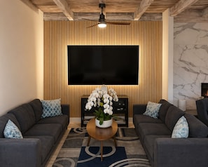 A modern living room with a large flat-screen TV, two sofas with patterned cushions, a wooden accent wall, and a marble fireplace.