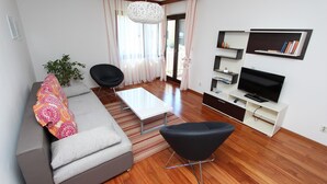 A modern living room with a grey sofa, black armchair, and a white coffee table. There is a TV on a white stand with shelves above it. - Apartment Mitton 2 (Rovinj)