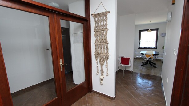A hallway with a wooden door, a macrame wall hanging, a dining area with a table and chairs, and a clock on the wall. - Apartment Mitton 2 (Rovinj)